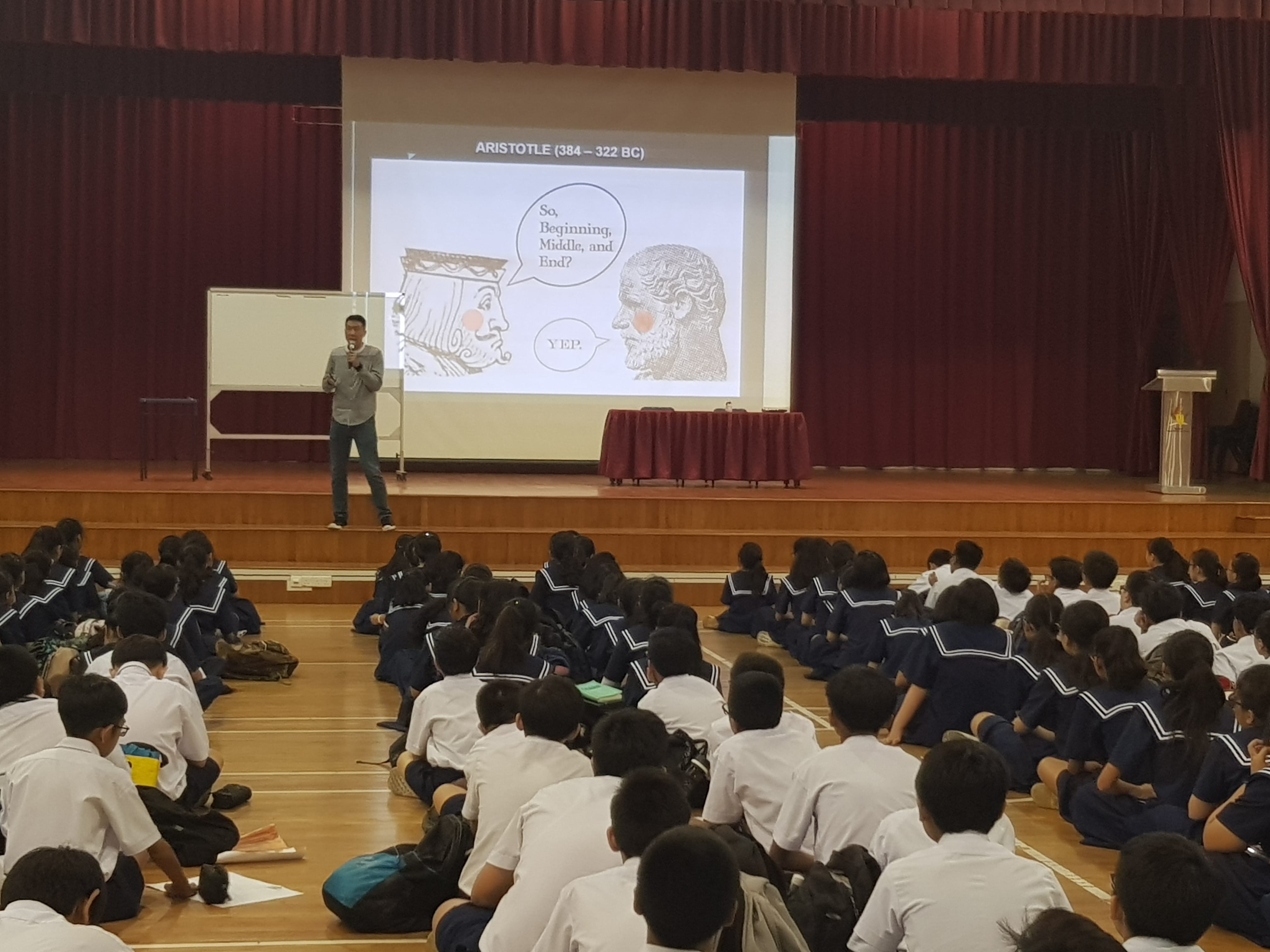 A speaker on stage presents an Aristotle slide to uniformed students seated on a school hall floor.